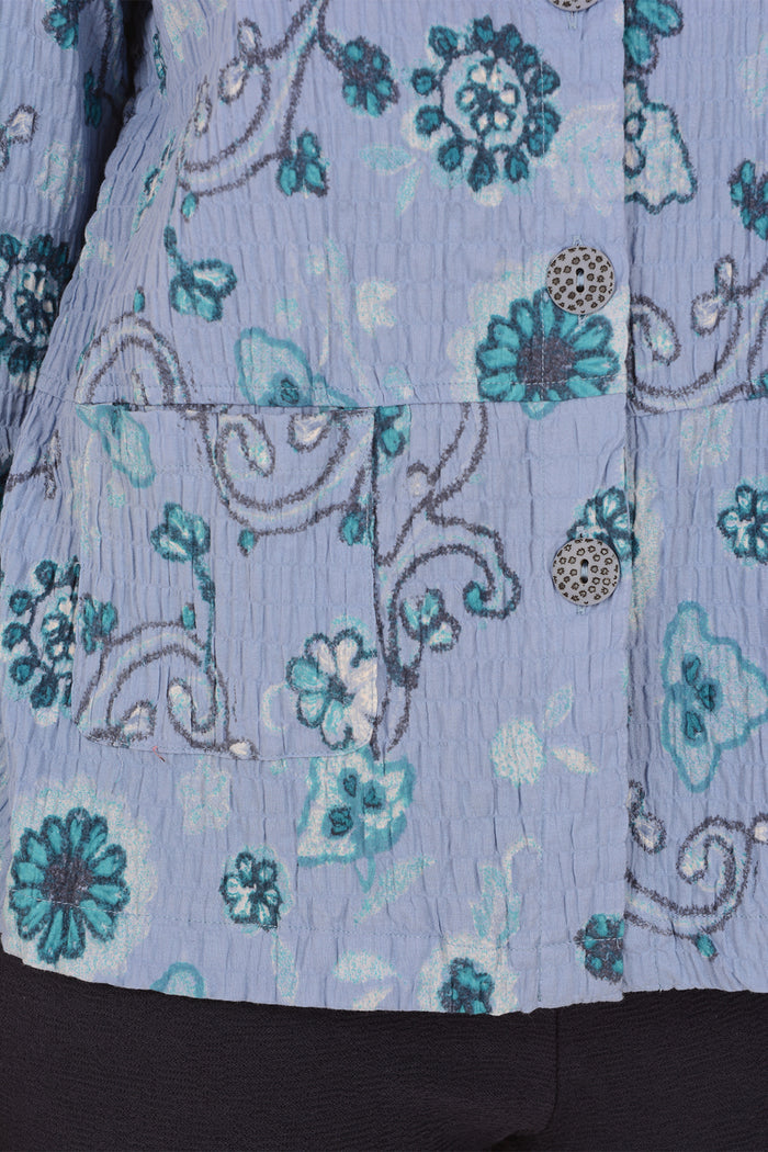 Close-up of a light blue crinkled fabric garment with a teal and white floral pattern, featuring decorative metallic buttons and a pocket seam.
