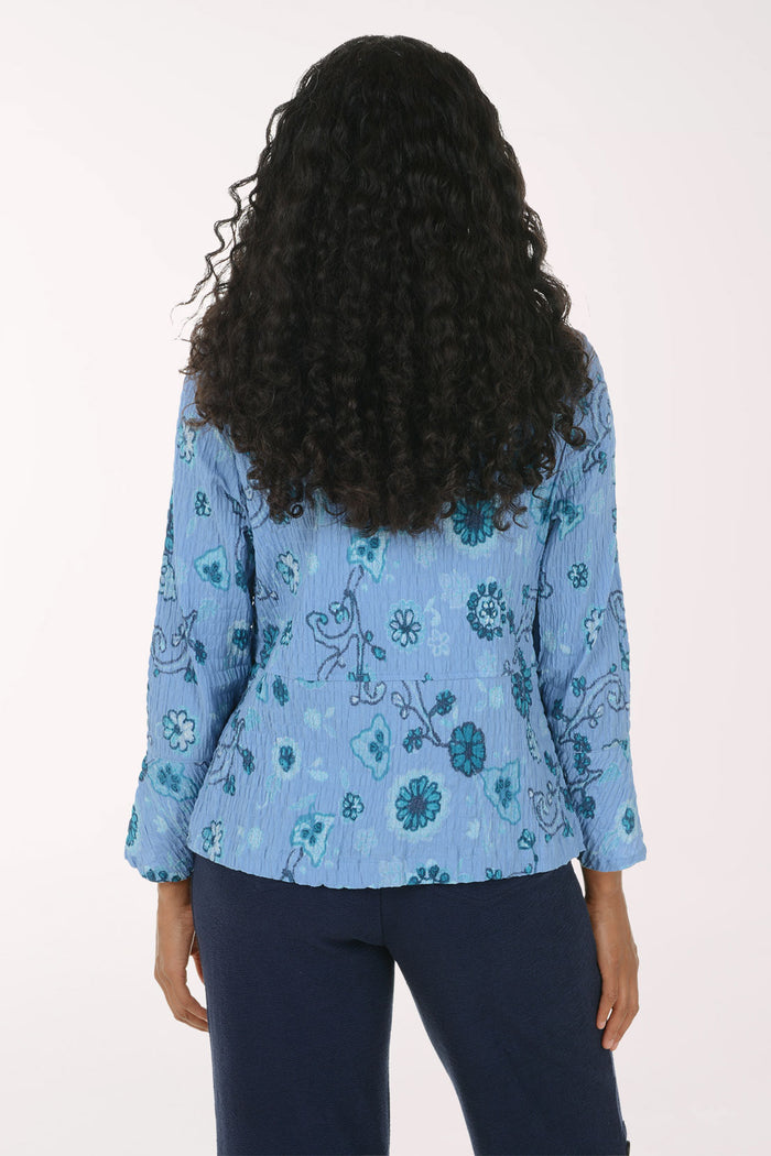 A woman viewed from the back wearing a light blue long-sleeve blouse with a teal and dark blue floral pattern, paired with dark navy pants, against a plain white background.