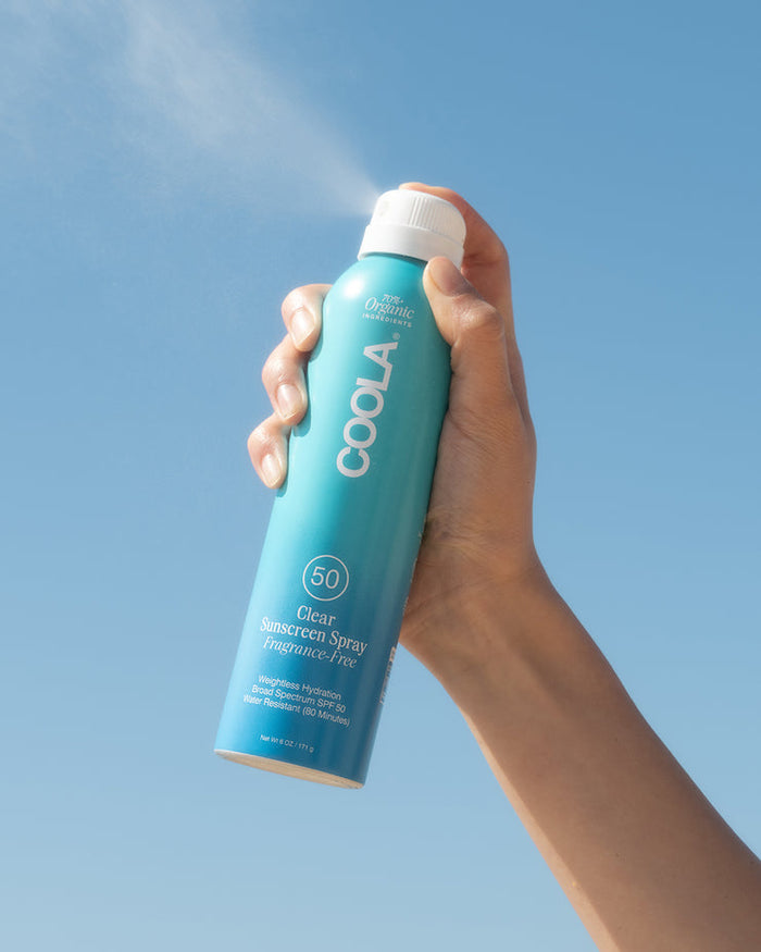 Hand holding Classic Body Organic Sunscreen Spray SPF 50 - 6 fl oz | COOLA, spraying clear sunscreen against a blue sky, promoting skincare and sun protection.