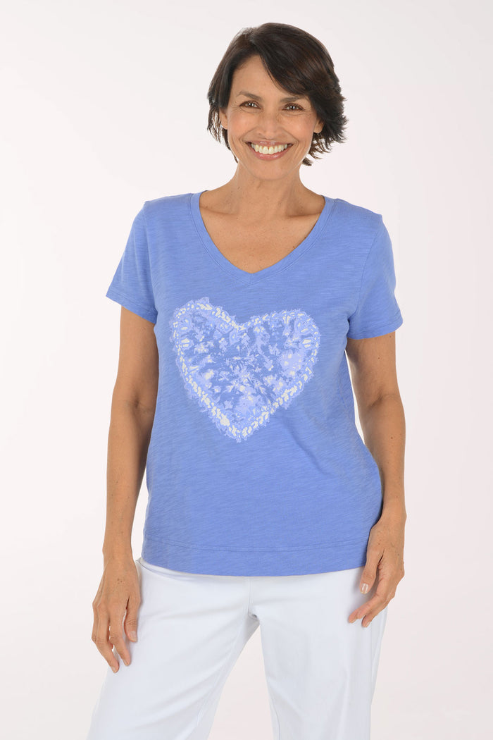 A smiling woman wearing a heathered periwinkle blue V-neck t-shirt with a white heart-shaped graphic on the front, paired with white pants, against a plain white studio background.