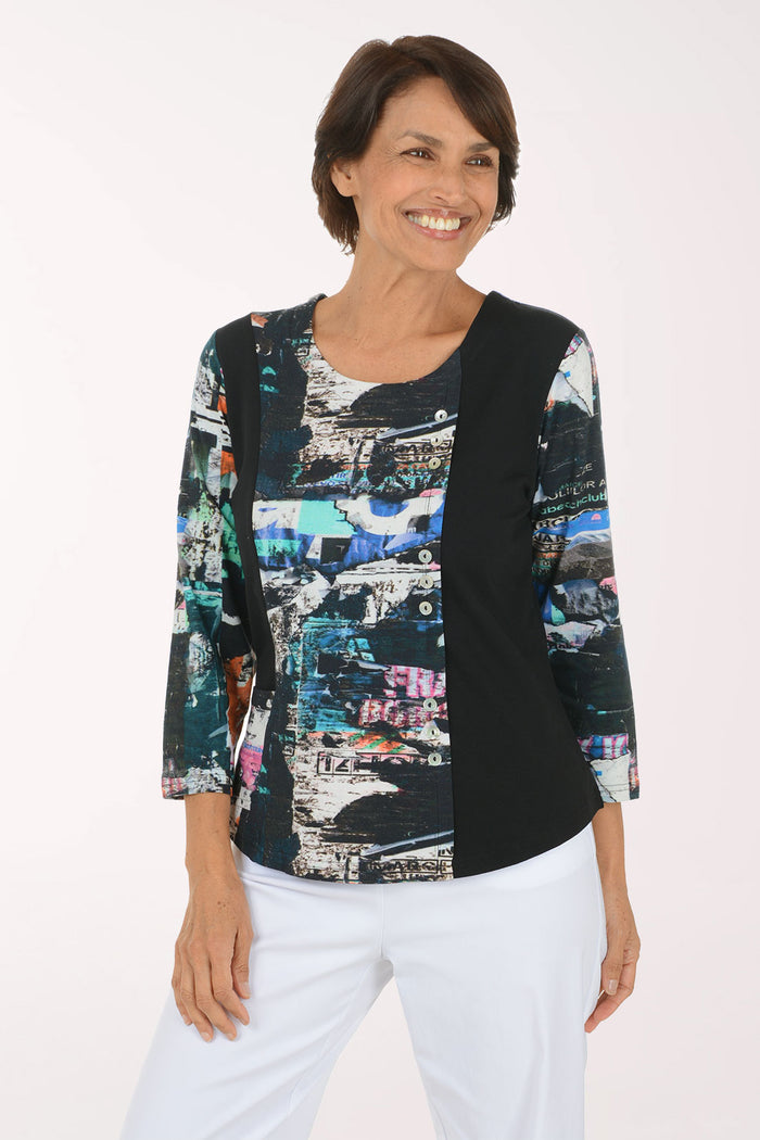 A woman modeling a three-quarter sleeve top with a black side panel and a multicolor abstract collage print on the front and sleeves, paired with white pants against a plain white background.