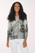 A woman with long, curly black hair is smiling and wearing a three-quarter sleeve top with an abstract, painterly print in gray, green, black, and red, paired with white pants, against a plain white studio background.