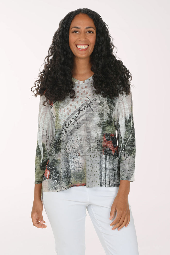 A woman with long, curly black hair is smiling and wearing a three-quarter sleeve top with an abstract, painterly print in gray, green, black, and red, paired with white pants, against a plain white studio background.
