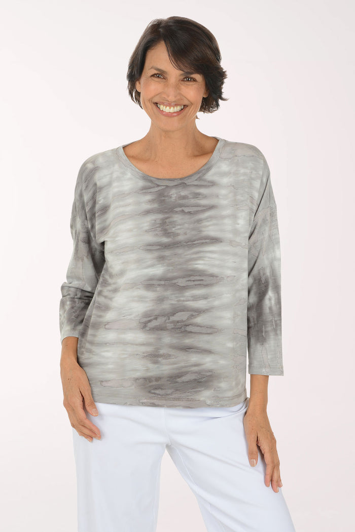 A smiling woman wearing a gray and white tie-dye long-sleeve top with white pants, standing against a plain light background.