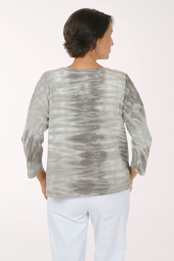 A smiling woman wearing a gray and white tie-dye long-sleeve top with white pants, standing against a plain light background.